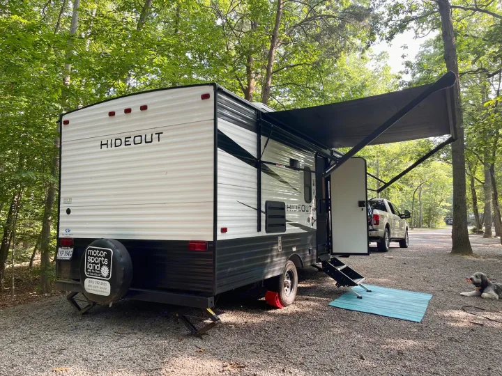 An RV rental, a white Hideout trailer, is parked on a wooded campsite with its awning extended and steps out. A dog relaxes on the gravel.. Keystone RV Hideout 2021