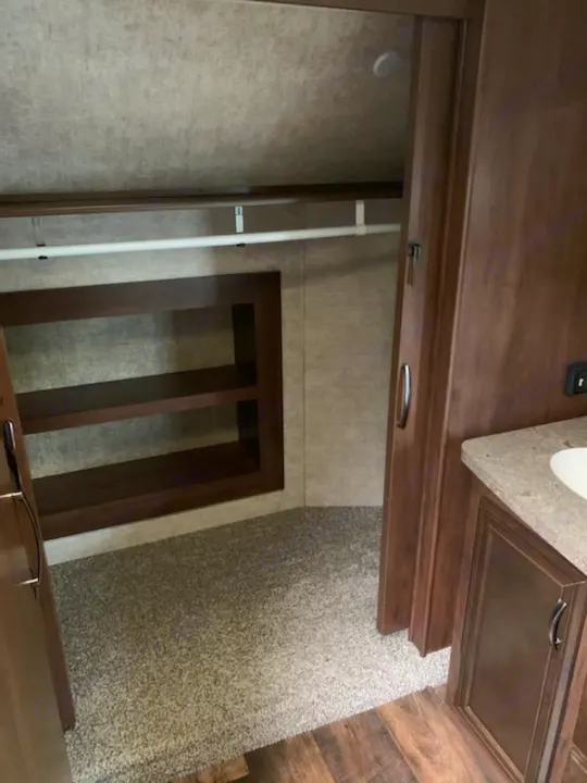 RV rental interior: compact closet with hanging rod, built-in shelves, and carpet, leading to a bathroom vanity area.. Forest River Sandpiper 2018