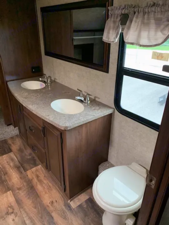 This RV rental bathroom boasts a modern double vanity with two sinks, a toilet, mirror, and wood-look flooring.. Forest River Sandpiper 2018