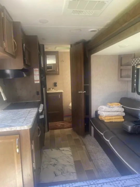 Inside an RV rental: compact kitchen, bathroom with sink/mirror, and living space with a black sofa & towels. Homey travel.. Dutchmen Coleman Light 2019