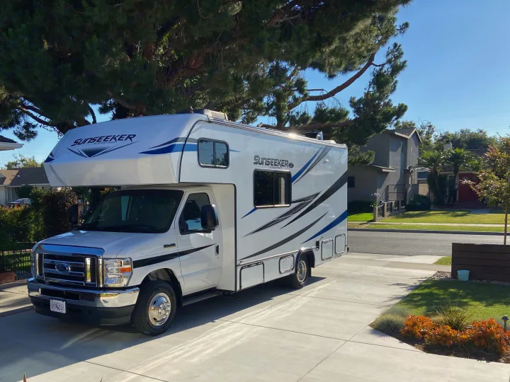 A white Sunseeker RV rental is parked on a sunny concrete driveway, with green trees overhead and houses in the background.. Forest River Sunseeker LE 2022