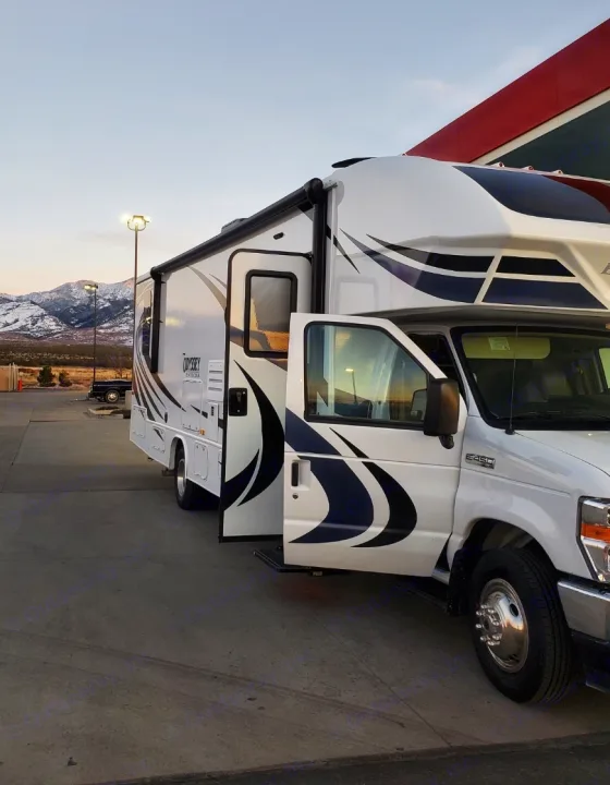 This white Class C RV rental, with its door open, is parked against a scenic backdrop of snowy mountains, ready for travel.. Entegra Coach Odyssey 2021