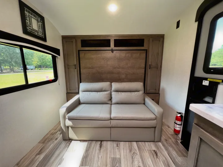 Comfortable RV rental interior featuring a sofa, convertible bed/murphy bed wall, and window with park view. Fire extinguisher.. Venture Rv Sonic 2023