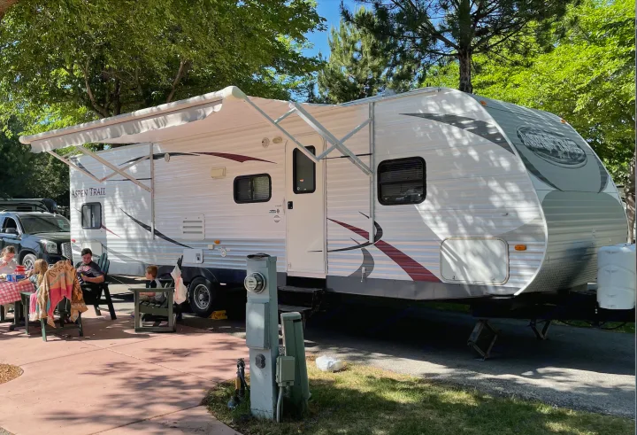 A family enjoys their RV rental travel trailer set up at a shaded campsite, relaxing outdoors under the extended awning.. Aspen Trail Aspen Trail Trailer 2013