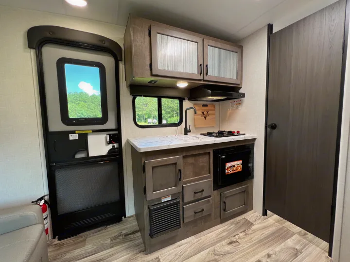 Modern RV rental kitchen with stove, microwave, cabinets. Includes entrance door, outdoor view, and fire extinguisher.. Venture Rv Sonic 2023