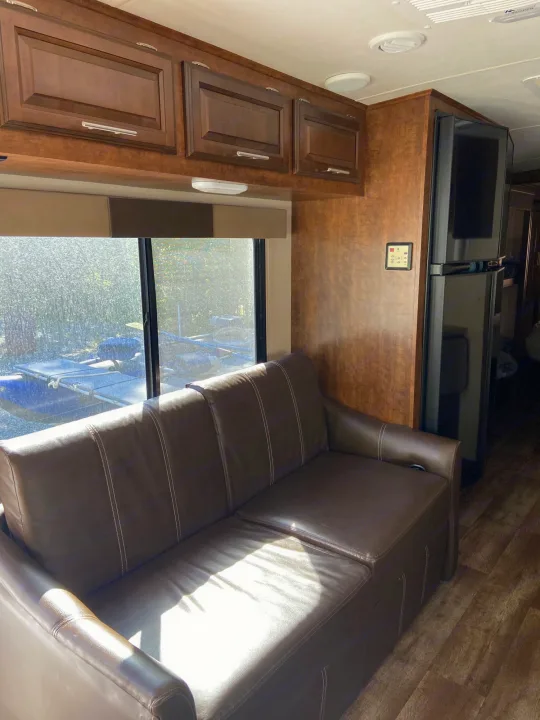 Cozy interior of an RV rental: brown leather sofa, overhead wood cabinets, and refrigerator by a large window with outdoor view.. Forest River Fr3 2017