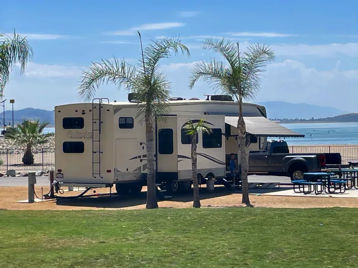 Sunny waterfront RV rental setup with palm trees, a large fifth-wheel, and a truck at a serene lake campsite.. Palomino Columbus 2018