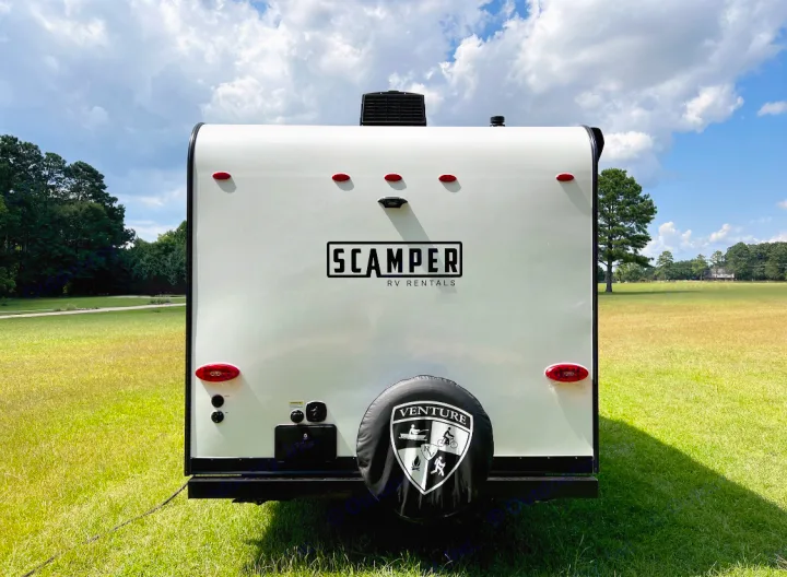 Rear view of a white Scamper RV rental with a 'Venture' spare tire, parked in a large green field under clouds.. Venture Rv Sonic 2023