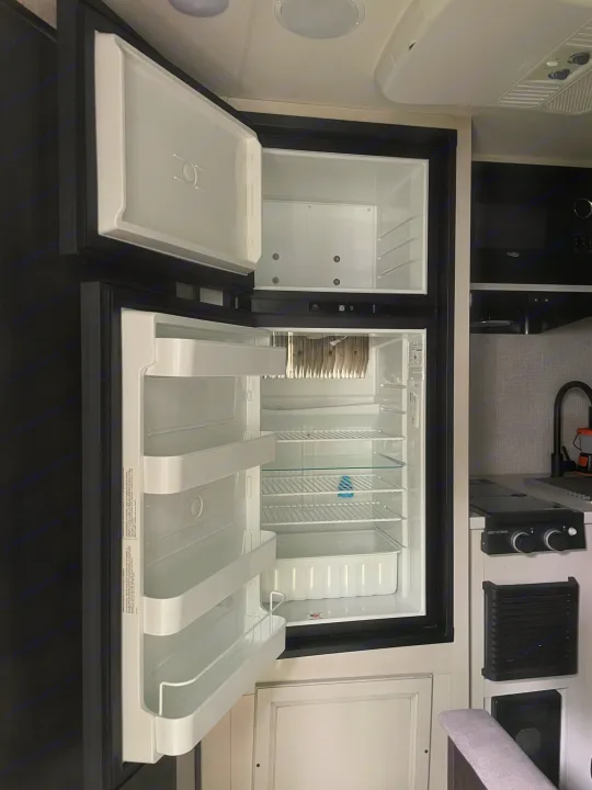 Inside an RV rental: A clean, empty refrigerator and freezer, with a partial view of the compact kitchen and stovetop.. Jayco Jay Flight SLX 2022