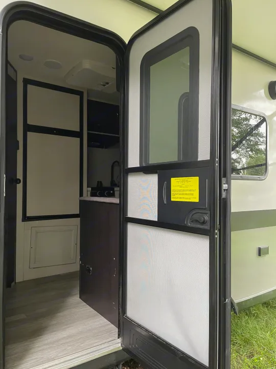 Open RV rental door showing the interior: compact kitchen area, refrigerator, and light wood-look flooring.. Jayco Jay Flight SLX 2022