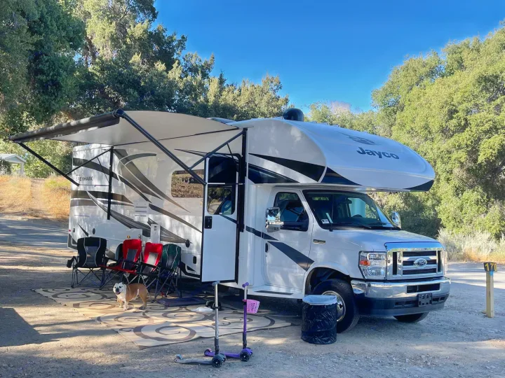 Relaxing at a sunny campsite. This white RV rental has an extended awning providing shade for a bulldog, chairs, and scooters.. Jayco Greyhawk 2022