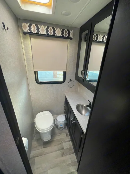 Bright RV bathroom: white toilet, vanity with sink, mirror, and window with patterned valance. Ideal for your RV rental.. Jayco Greyhawk 2022