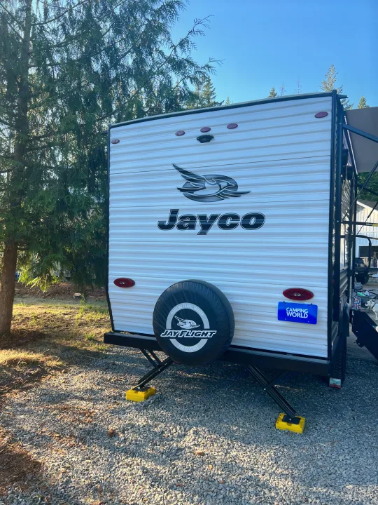 Rear view of a white Jayco travel trailer parked on gravel at a sunny campsite with stabilizing jacks. Great for an RV rental!. Jayco Jay Flight SLX Baja Edition 2023