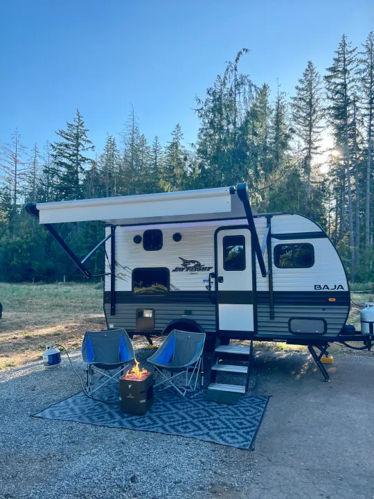 This RV rental is ready for adventure! A cozy Jay Flight travel trailer setup with chairs & fire pit in a sunny forest camp.. Jayco Jay Flight SLX Baja Edition 2023