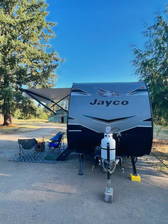 Jayco RV rental travel trailer at a sunny campsite, featuring an extended awning, chairs, and a warm fire pit.. Jayco Jay Flight SLX Baja Edition 2023