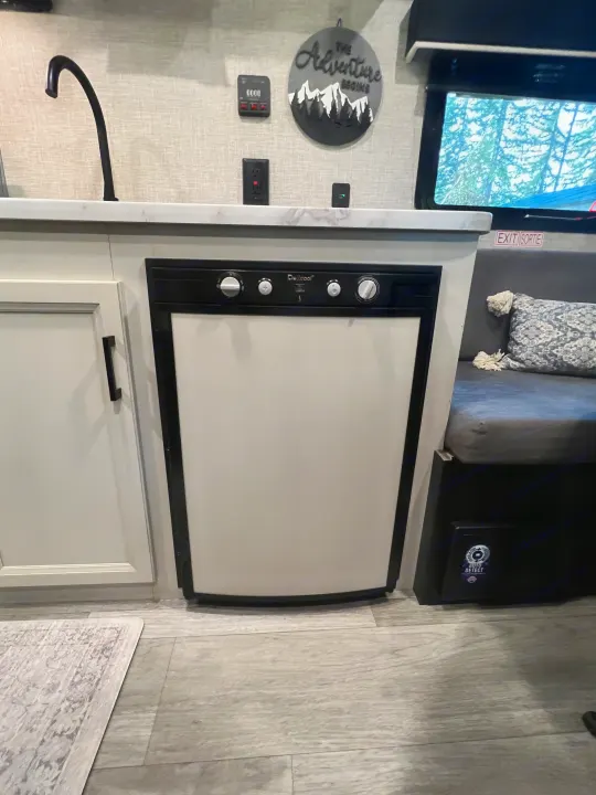 Inside an RV rental: a compact kitchen with a black faucet, Delcool mini-fridge, and dinette with "The Adventure Begins" decor.. Jayco Jay Flight SLX Baja Edition 2023