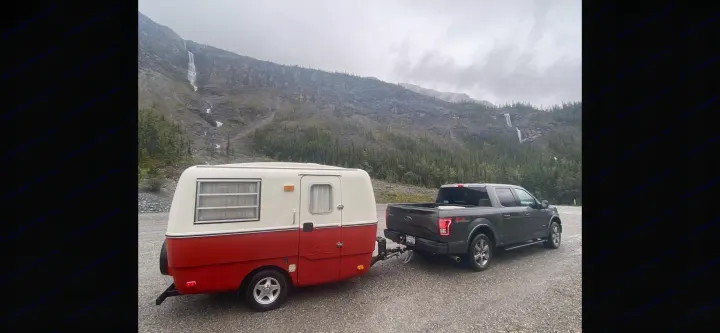 RV rental: A vintage travel trailer towed by a truck, parked amidst mountains with waterfalls under an overcast sky.. Trillium Rv other 1975