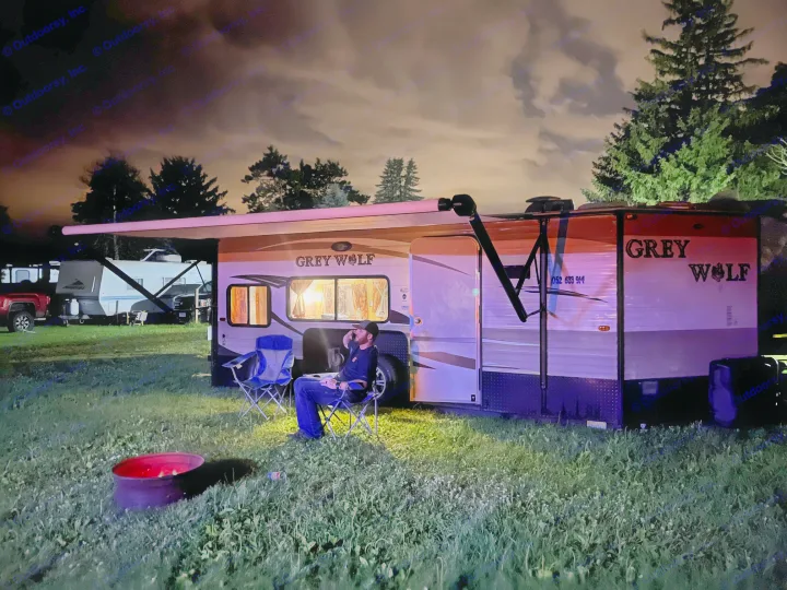 A man enjoys a cozy night by a fire pit outside his Grey Wolf RV rental, under a lit awning.. Forest River Cherokee Grey Wolf 2015