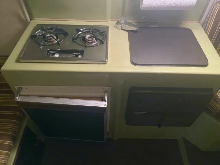 Retro RV rental kitchen: two-burner stove, covered sink, fridge, and heater in a green camper interior.. Trillium Rv other 1975