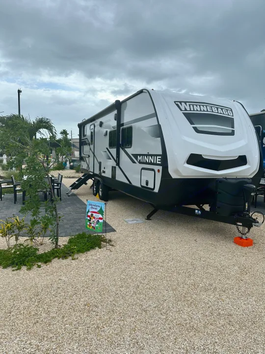 White Winnebago Minnie travel trailer RV rental at a spacious gravel campsite with a patio & outdoor seating under an overcast sky.. Winnebago Minnie 2301BH 2023