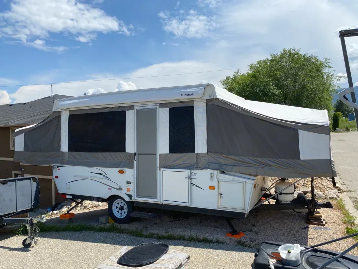 Grey & white pop-up RV rental with extended awning, parked on gravel under a blue, cloudy sky for camping.. Palomino Yearling 2012
