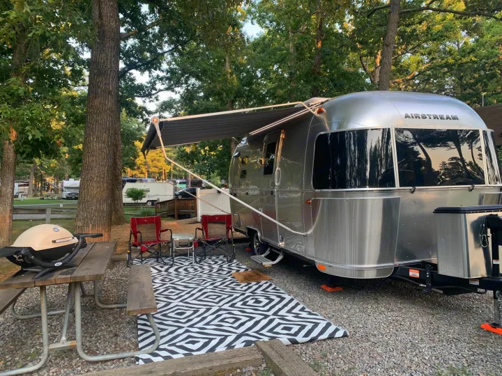 An Airstream RV rental is set up at a wooded campsite with an awning, outdoor rug, and picnic table with a grill.. Airstream Caraval 2021