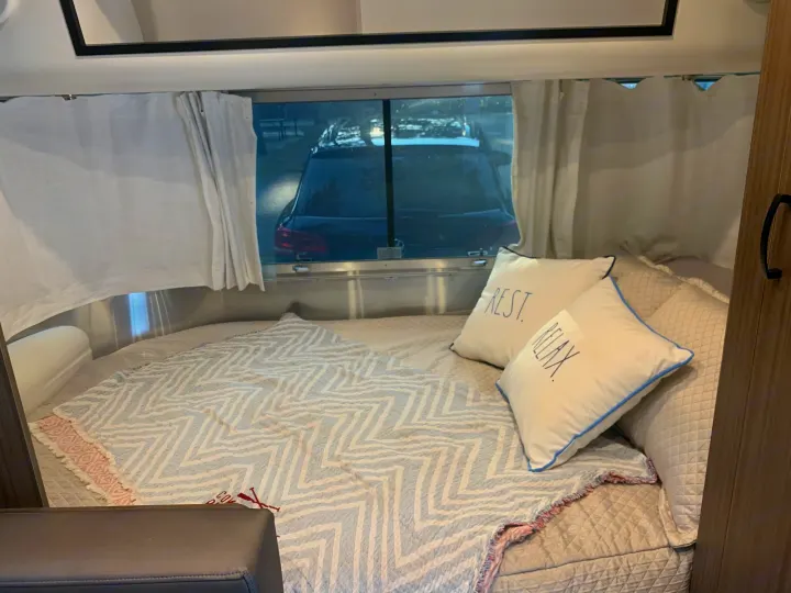 Comfortable RV rental bedroom with a cozy bed, "REST" & "RELAX" pillows, and a window view of a parked car.. Airstream Caraval 2021