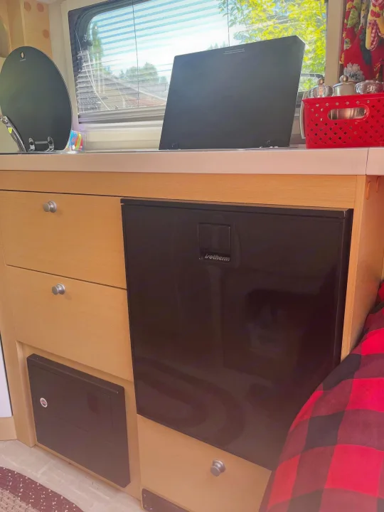 RV rental's compact kitchen area with an Isotherm fridge, wood drawers, and counter by a window.. nuCamp T@B 2020
