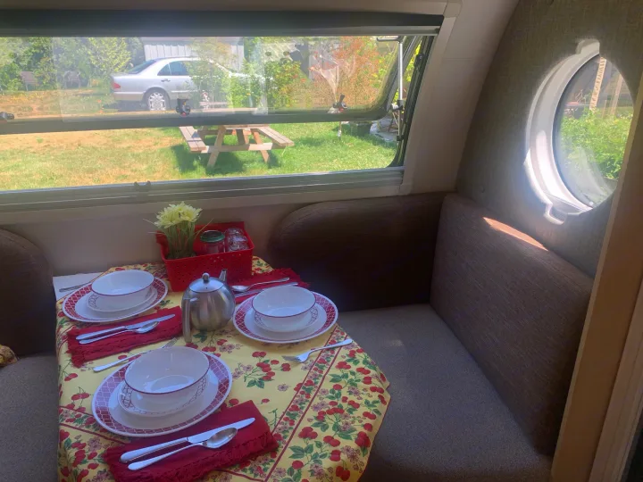Cozy RV rental dining area, table set for two. Large windows offer a bright view of a yard with a car and picnic table.. nuCamp T@B 2020
