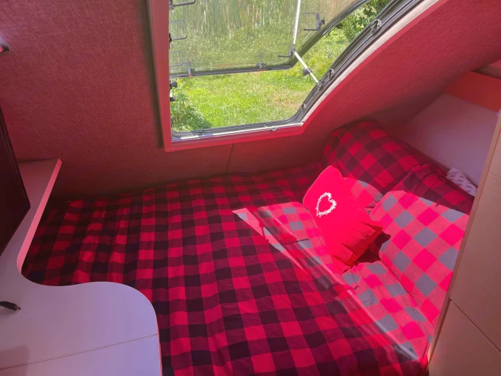 Cozy RV rental interior with a comfy red plaid bed, heart pillow, and open window showing green nature.. nuCamp T@B 2020