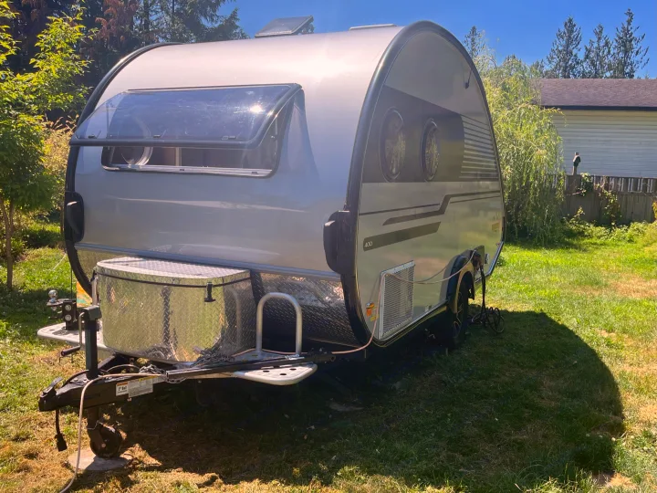 This modern silver teardrop RV rental, a compact travel trailer, is parked on a sunny green lawn.. nuCamp T@B 2020