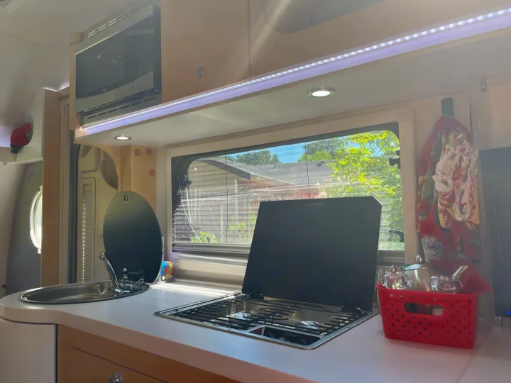 Inside an RV rental: a bright kitchen area with a stainless steel sink, covered Dometic stove, and an overhead microwave.. nuCamp T@B 2020