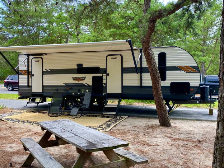 Scenic RV rental setup: a large travel trailer with awning extended, chairs, and picnic table at a sandy campsite.. Forest River Wildwood 2021