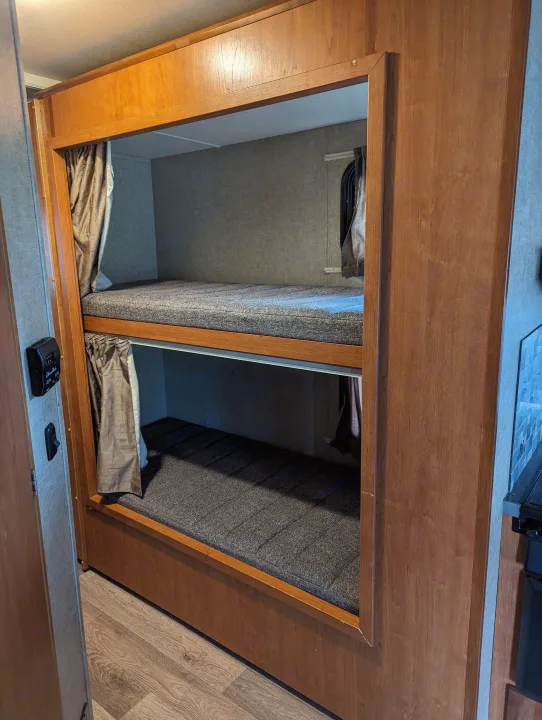 Stacked bunk beds with grey mattresses and privacy curtains inside an RV rental, featuring warm wood trim.. Winnebago Spirit 2019