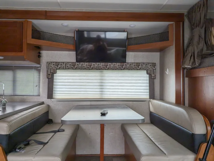 Inside an RV rental, showing the dinette area with light-colored booth seating, a table, TV, and window blinds.. Winnebago Spirit 2019