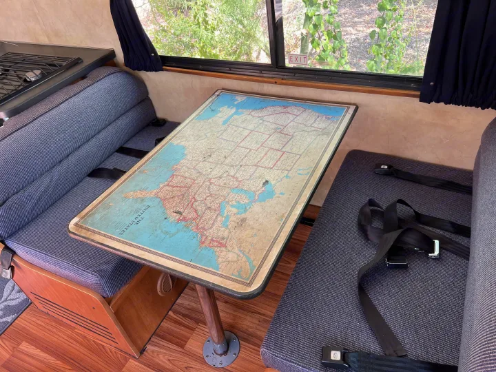 Inside an RV rental, a dining area features a large map table, blue benches with seatbelts, and a window looking out to nature.. Thor Majestic 2013
