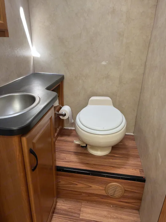 Inside an RV rental: a practical bathroom with beige toilet, stainless steel sink, wood-look floor, and sunlight.. Thor Majestic 2013