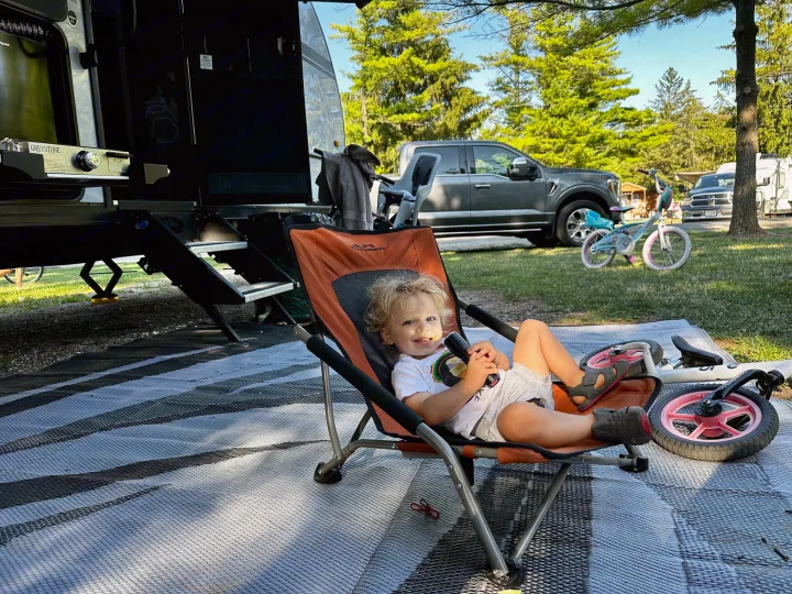 RV rental camping trip: A joyful toddler relaxes in a chair on a mat, with bikes & RVs under sunny trees.. Forest River Cherokee Grey Wolf Black Label 2021