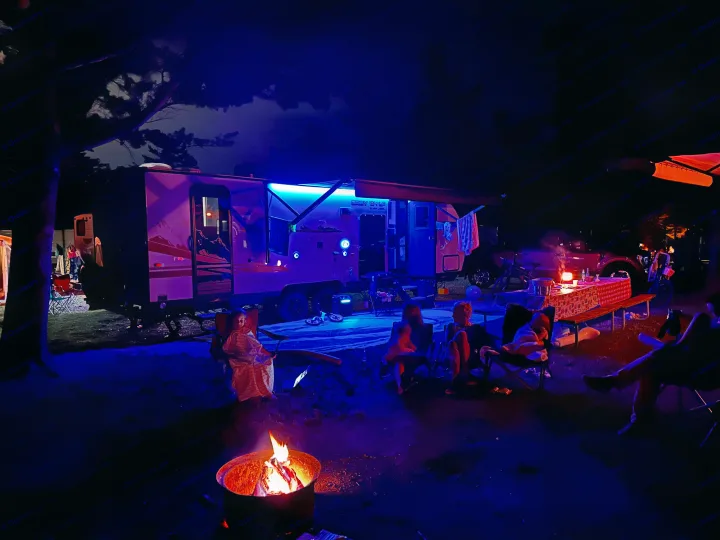 Enjoying a cozy night at the campsite with an RV rental. People relax by a warm fire pit, bathed in the RV's ambient blue light.. Forest River Cherokee Grey Wolf Black Label 2021