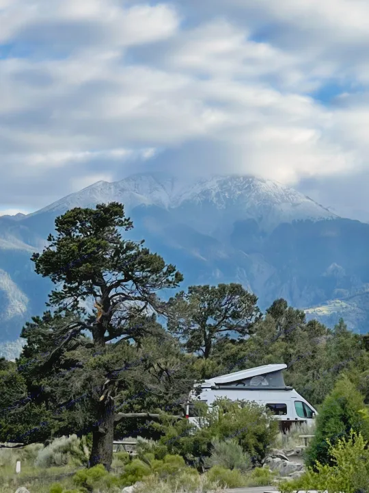 A white RV rental rests among green trees with a majestic, snow-capped mountain behind, under a cloudy sky. Ideal for nature adventures.. Winnebago Solis 2021