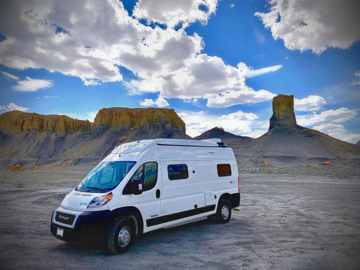 White RV rental parked in a vast desert landscape with striking rock formations under a cloudy blue sky, suggesting adventure.. Winnebago Solis 2021
