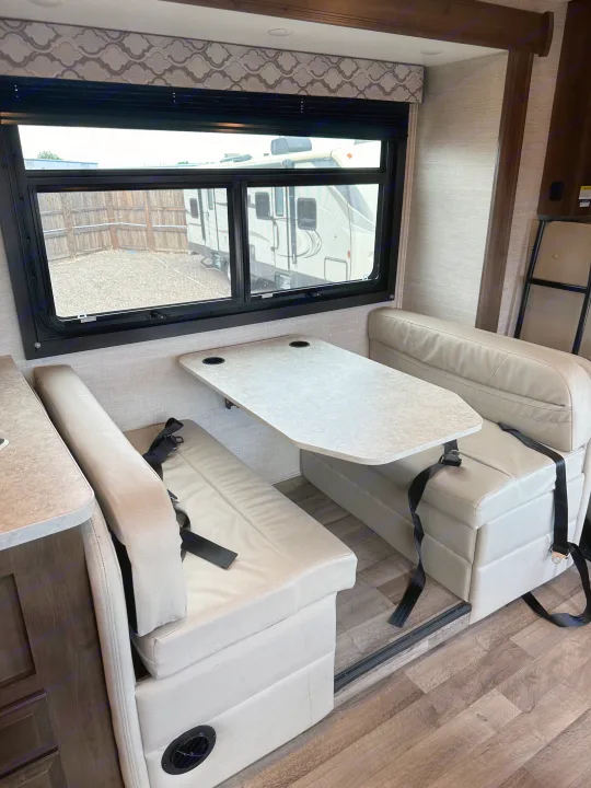 RV rental dinette interior: Light-toned booth seating with seatbelts, a table with cupholders, and a large window view.. Jayco Redhawk 2021
