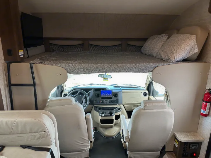Inside an RV rental: cockpit with steering wheel & screen, front seats, and an overcab bunk bed with pillows, TV, and ladder.. Jayco Redhawk 2021