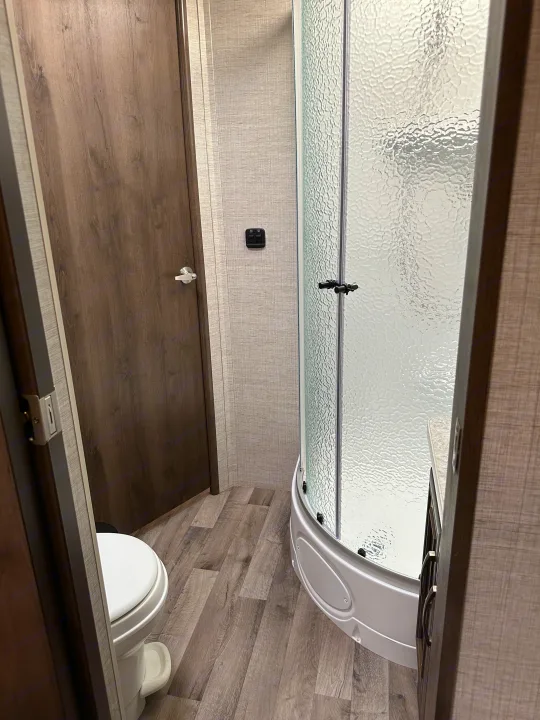 Step inside this RV rental's compact bathroom, featuring a wood door, curved glass shower, and partially visible toilet.. Jayco Redhawk 2021