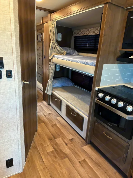 RV rental featuring two cozy bunk beds with light bedding, a compact kitchen with a stove, and wood-look flooring.. Jayco Redhawk 2021