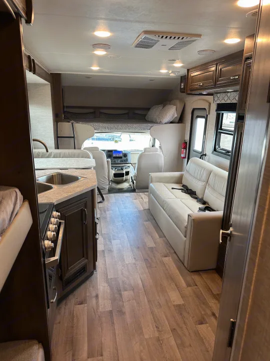 Modern RV rental interior: kitchen with sink/stove, sofa, driver's cabin, and overhead bunk. Brightly lit living space.. Jayco Redhawk 2021
