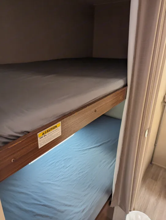 Inside an RV rental, bunk beds are made with gray and blue sheets. A caution sign on the top bunk warns of a 300 lbs weight limit.. Dutchmen Coleman Lantern LT 2017