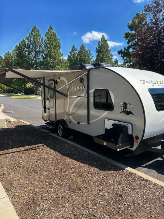 This light-colored r-pod travel trailer, an RV rental, is parked with its awning extended & pull-out grill. Sunny day, trees in background.. R-Pod Hood River Edition 2020