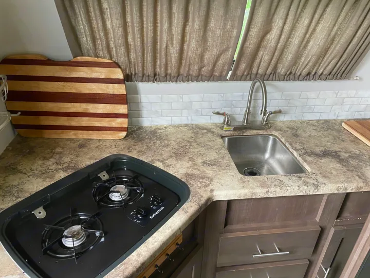 RV rental kitchen: two-burner stove, stainless steel sink, mottled counter, white backsplash, and brown cabinets/curtains.. R-Pod R-Pod Trailer 2017