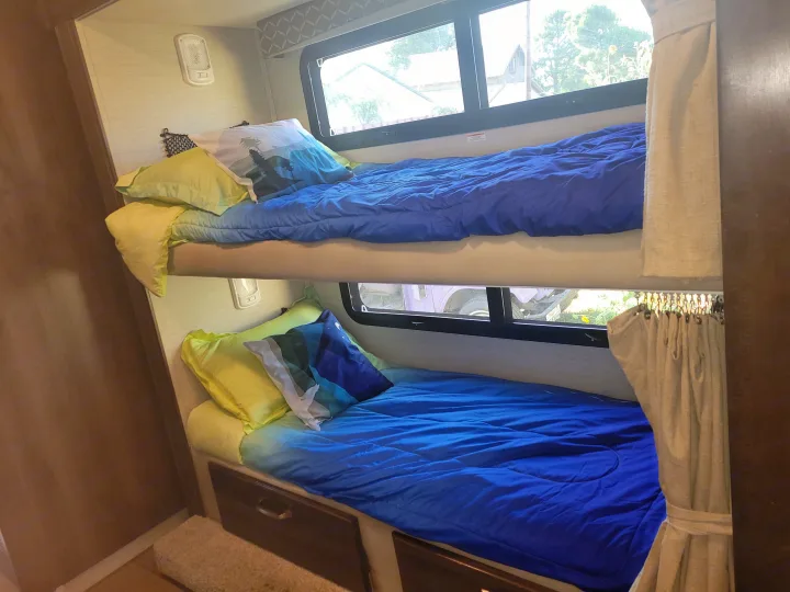 Inside an RV rental, two bunk beds with blue comforters & decorative pillows offer comfy sleeping, with windows & storage drawers visible.. Jayco Greyhawk 2018